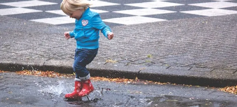Boy in Blue Jacket Hopping on Water Puddle